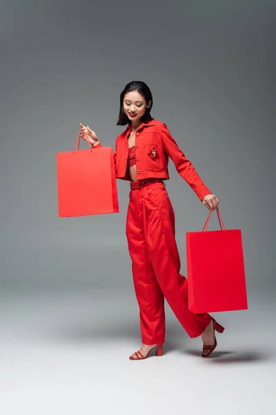 Longitud completa de sonriente mujer asiática en traje rojo y sandalias de tacón posando con bolsas de compras sobre fondo gris - foto de stock