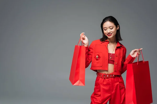 Felice donna asiatica in abiti eleganti in posa con borse della spesa rosse isolate sul grigio — Foto stock