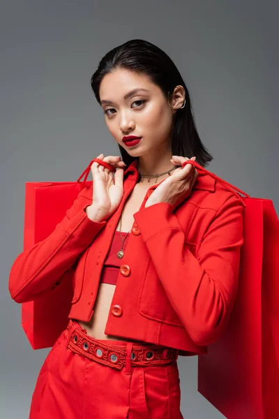 Красивая азиатская женщина в модном пиджаке с красными сумками для покупок и глядя на камеру, изолированную на сером — стоковое фото
