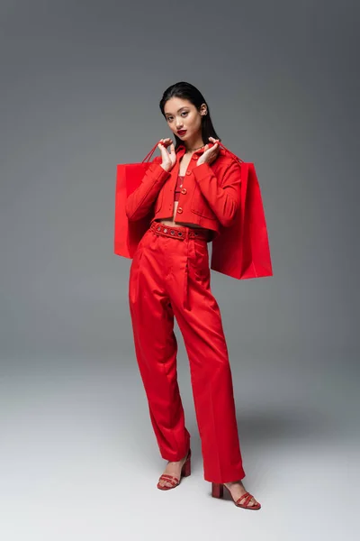 Volle Länge der asiatischen Frau in rotem Outfit mit Einkaufstaschen und Blick in die Kamera auf grauem Hintergrund — Stockfoto