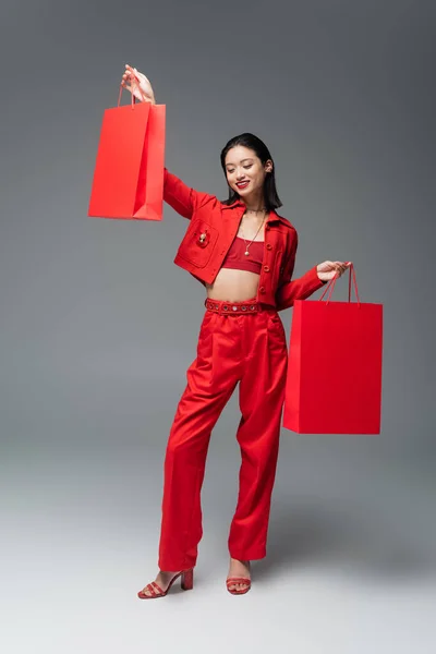 Full length of cheerful asian woman in trendy attire posing with red shopping bags on grey background — Stock Photo