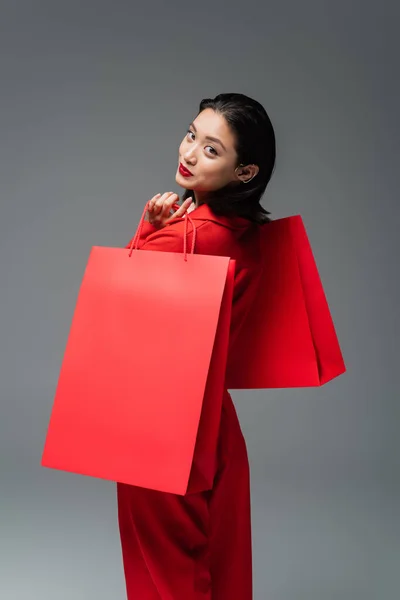 Morena asiática mujer con rojo compras bolsas sonriendo a cámara aislada en gris - foto de stock