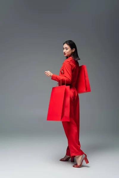 Полная длина модной азиатской женщины с красными сумками покупки глядя в сторону на сером фоне — стоковое фото