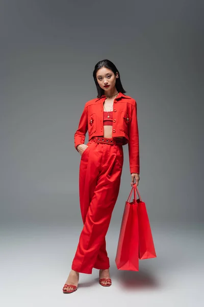 Volle Länge der stilvollen asiatischen Frau posiert mit Einkaufstaschen und Hand in Tasche der roten Hose auf grauem Hintergrund — Stockfoto