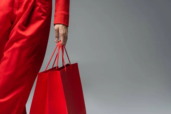 Vista parcial de la mujer en pantalones rojos de pie con bolsa de compras sobre fondo gris - foto de stock