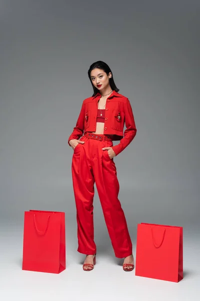Young asian model in red jacket and trousers posing with hands in pockets near shopping bags on grey background — Stock Photo