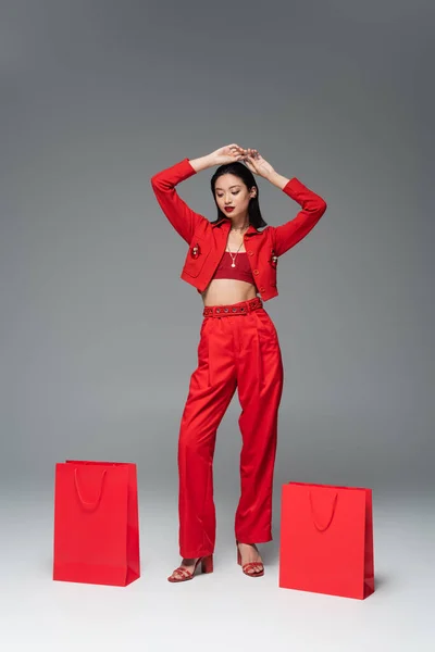 Piena lunghezza di donna asiatica in abito elegante posa con le mani sopra la testa vicino borse della spesa rosse su sfondo grigio — Foto stock