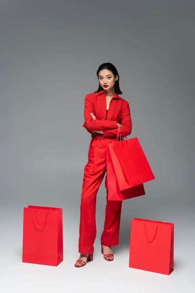 Повна довжина азіатської жінки в червоному стильному костюмі, що стоїть з схрещеними руками біля сумки для покупок на сірому фоні — стокове фото