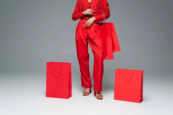 Частичный вид женщины в брюках и сандалиях, стоящей рядом с красными пакетами покупок на сером фоне — стоковое фото