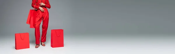 Vista ritagliata della donna in abito alla moda in posa vicino a borse della spesa rosse su sfondo grigio, banner — Foto stock