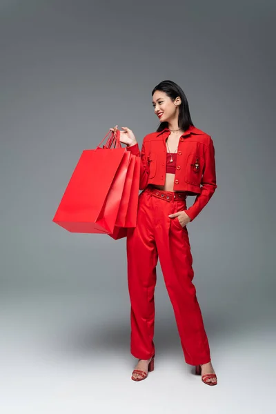 Longitud completa de mujer asiática en traje elegante rojo posando con bolsas de compras y mano en bolsillo sobre fondo gris - foto de stock