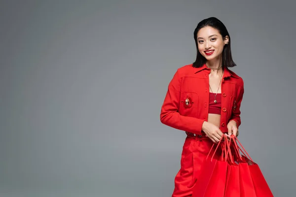 Fashionable asian woman smiling at camera while standing with red shopping bags isolated on grey — Stock Photo