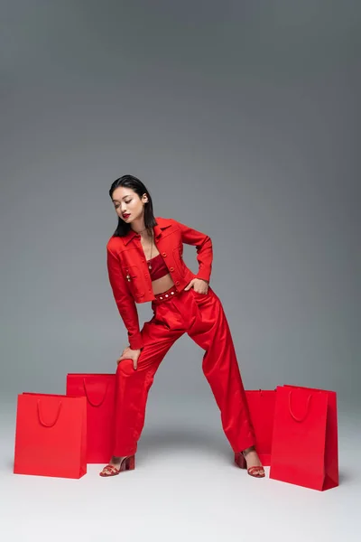 Asiatin in voller Länge im roten schicken Anzug posiert mit der Hand in der Tasche in der Nähe von Einkaufstaschen auf grauem Hintergrund — Stockfoto