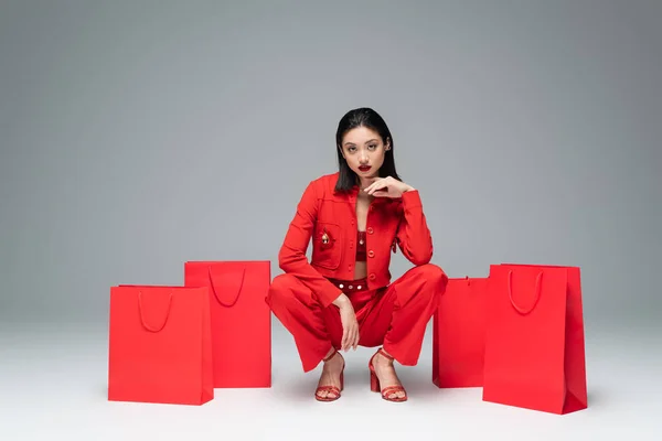 Trendy asian woman in red sitting on haunches near shopping bags and looking at camera on grey background — Stock Photo