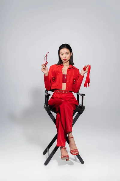 Longitud completa de elegante mujer asiática en traje rojo sosteniendo guante y gafas de sol mientras está sentado sobre fondo gris - foto de stock