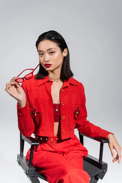 Брюнетка азиатка в красной одежде сидит на стуле в модных солнцезащитных очках, изолированных на сером — стоковое фото