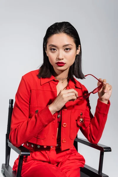 Young asian woman with red lips holding stylish sunglasses while sitting isolated on grey — Stock Photo