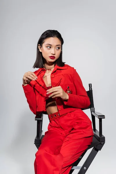 Asian woman in red elegant suit sitting on chair and holding sunglasses while looking away isolated on grey — Stock Photo