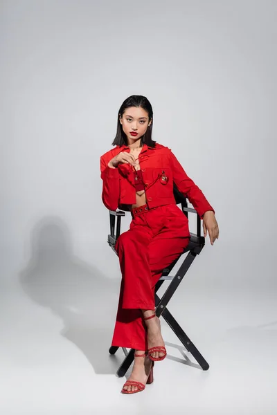 Comprimento total do modelo asiático em jaqueta vermelha e calças segurando óculos de sol elegantes enquanto sentado na cadeira no fundo cinza — Fotografia de Stock