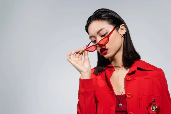 Young asian woman in red jacket holding stylish sunglasses isolated on grey — Stock Photo