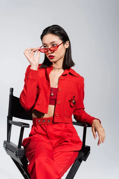 Elegante asiático mujer en rojo ropa celebración gafas de sol y mirando hacia otro lado mientras sentado en silla aislado en gris - foto de stock