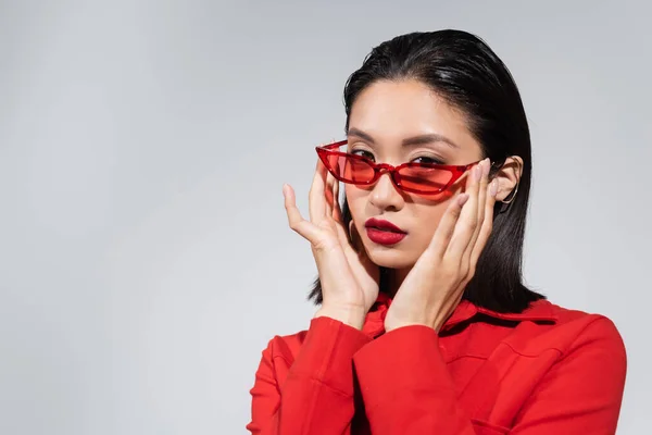 Portrait of young asian woman adjusting red and stylish sunglasses while looking at camera isolated on grey — Stock Photo