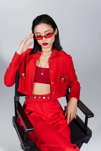Trendige asiatische Frau in roter Kleidung, die Sonnenbrille anpasst, während sie isoliert auf einem Stuhl in grau sitzt — Stockfoto