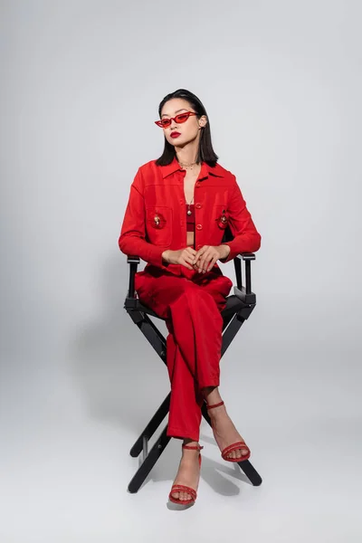 Asiatisches Model in voller Länge im roten eleganten Outfit und Sonnenbrille auf Stuhl auf grauem Hintergrund sitzend — Stockfoto