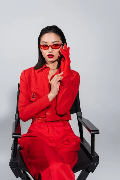 Mujer asiática de moda en traje elegante y gafas de sol con guante rojo aislado en gris - foto de stock