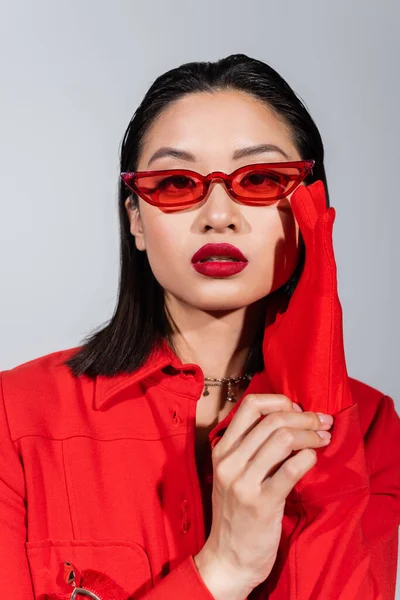 Портрет азиатки в красной куртке и перчатке, регулирующей модные солнцезащитные очки, изолированные на сером — стоковое фото