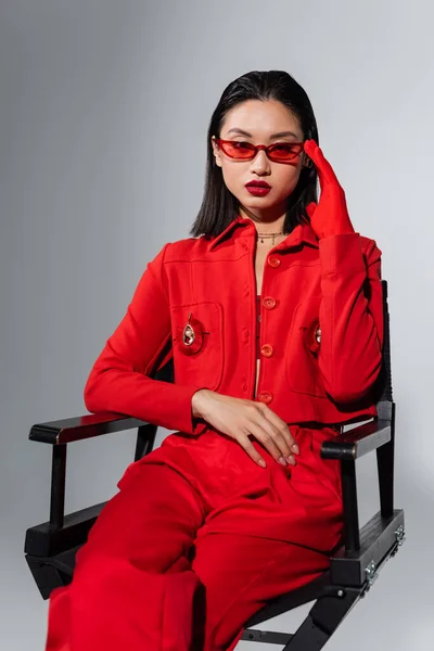 Asiatico modello in rosso vestito toccare elegante occhiali da sole mentre seduto isolato su grigio — Foto stock