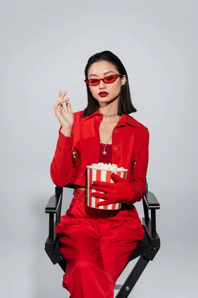 Брюнетка азіатська жінка в стильних сонцезахисних окулярах і червоний костюм сидить з відром попкорну ізольовані на сірому — стокове фото