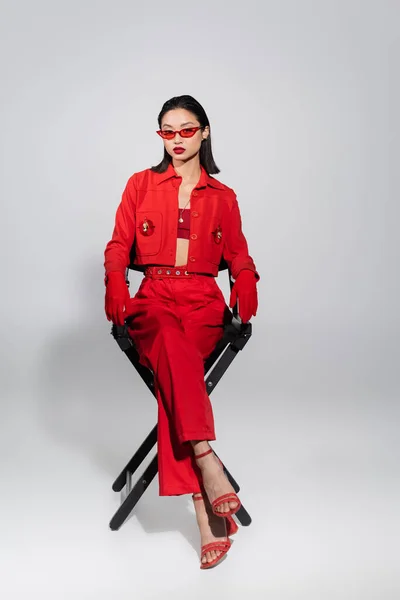 Pleine longueur de femme asiatique en rouge lunettes de soleil élégantes et costume élégant assis sur la chaise sur fond gris — Photo de stock