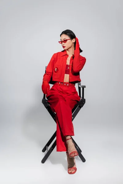 Full length of asian model in red outfit and sunglasses sitting on chair and looking away on grey background — Stock Photo