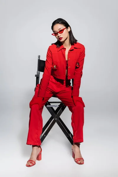 Повна довжина модної азіатки в червоному костюмі і сонцезахисних окулярах позує на стільці на сірому фоні — стокове фото