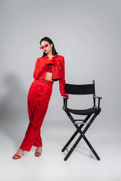 Full length of brunette asian woman in red suit and sunglasses posing near chair on grey background — Stock Photo