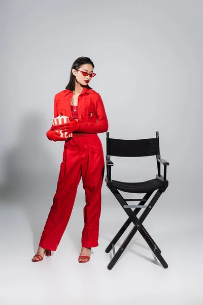 Full length of asian woman in red and stylish outfit standing with bucket of popcorn near chair on grey background — Stock Photo
