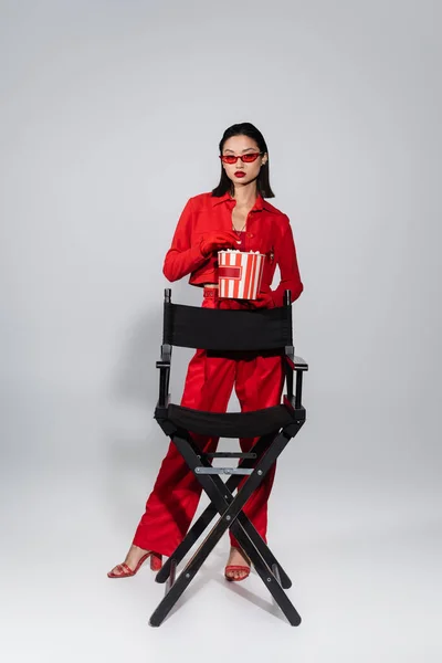 Mode asiatique femme en rouge lunettes de soleil tenant seau de pop-corn près de chaise sur fond gris — Photo de stock