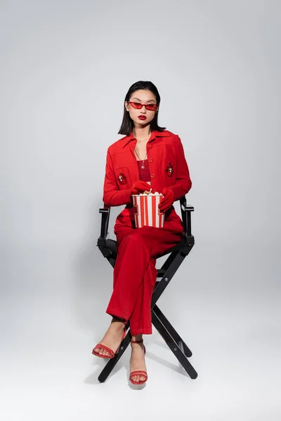 Full length of asian woman in trendy sunglasses and red elegant suit sitting with popcorn bucket on grey background — Stock Photo