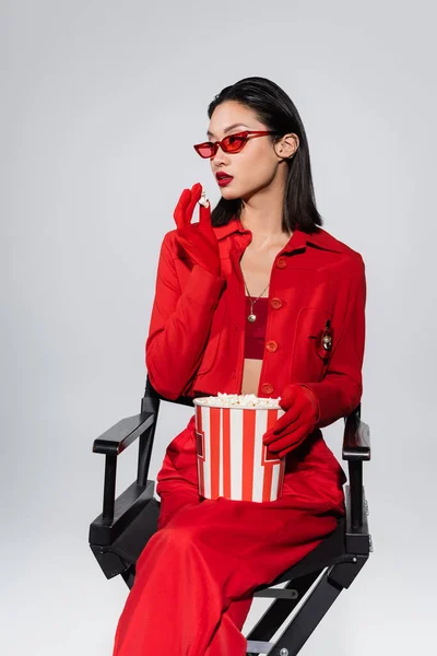 Модная азиатка в модных солнцезащитных очках и красной куртке держит попкорн и смотрит в сторону изолированной на сером — стоковое фото