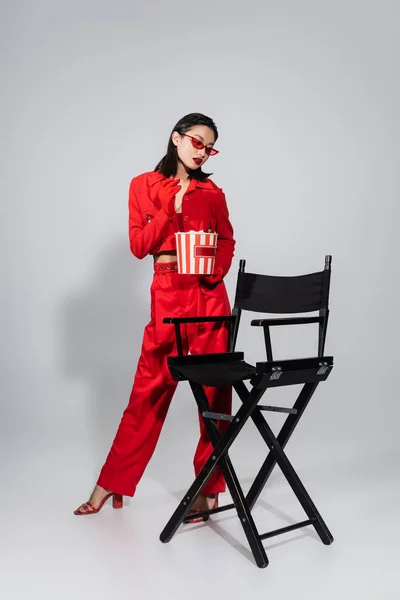 Piena lunghezza di bruna donna asiatica in occhiali da sole alla moda e abito rosso in piedi con popcorn vicino sedia su sfondo grigio — Foto stock
