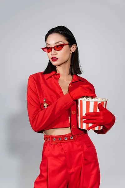 Азиатская женщина в модных солнцезащитных очках и красном наряде держит ведро попкорна и смотрит в сторону на сером фоне — стоковое фото
