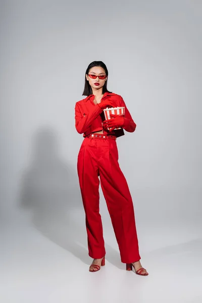 Full length of asian woman wearing sunglasses and red suit with gloves holding popcorn bucket on grey background — Stock Photo