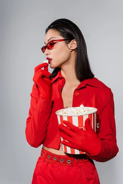 Morena mujer asiática en ropa roja y de moda y gafas de sol comiendo palomitas de maíz aisladas en gris - foto de stock