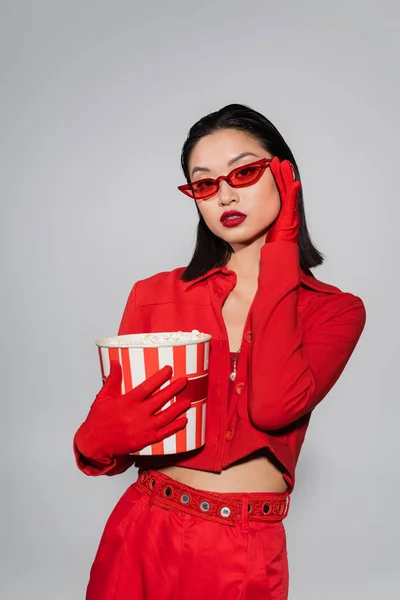 Азиатка в красных перчатках держит попкорн и регулирует модные солнцезащитные очки, изолированные на сером — стоковое фото