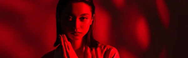 Портрет азіатки з молитовними руками дивиться на камеру на темному тлі з червоним світлом, банер — стокове фото