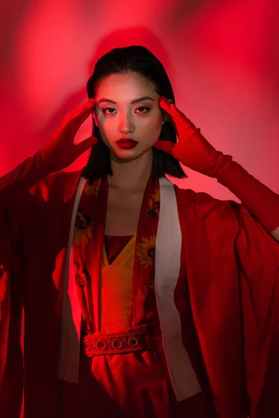 Young asian woman in kimono cape and gloves posing with hands near face on abstract background with red light — Stock Photo
