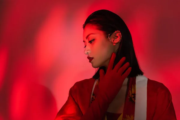 Young asian woman with makeup and ear cuff holding hand in glove near neck on shaded background with red light — Stock Photo