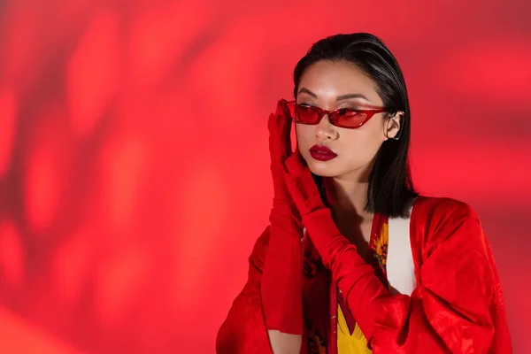 Trendy asian woman in red gloves and trendy sunglasses holding hands near face on abstract red background — Stock Photo