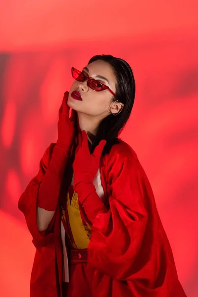 Sensual asian woman in gloves and sunglasses looking at camera on coral red background with shadow — Stock Photo